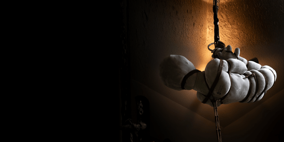 A shibari practice doll suspended in rope, lit from one side in a dark room