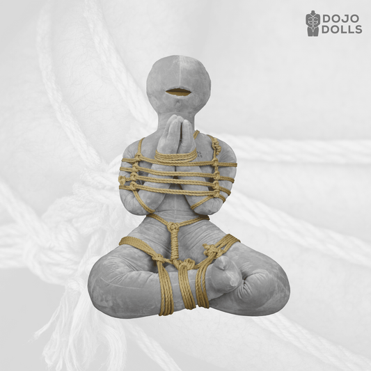 A shibari practice doll sitting cross-legged in a chest harness of jute rope